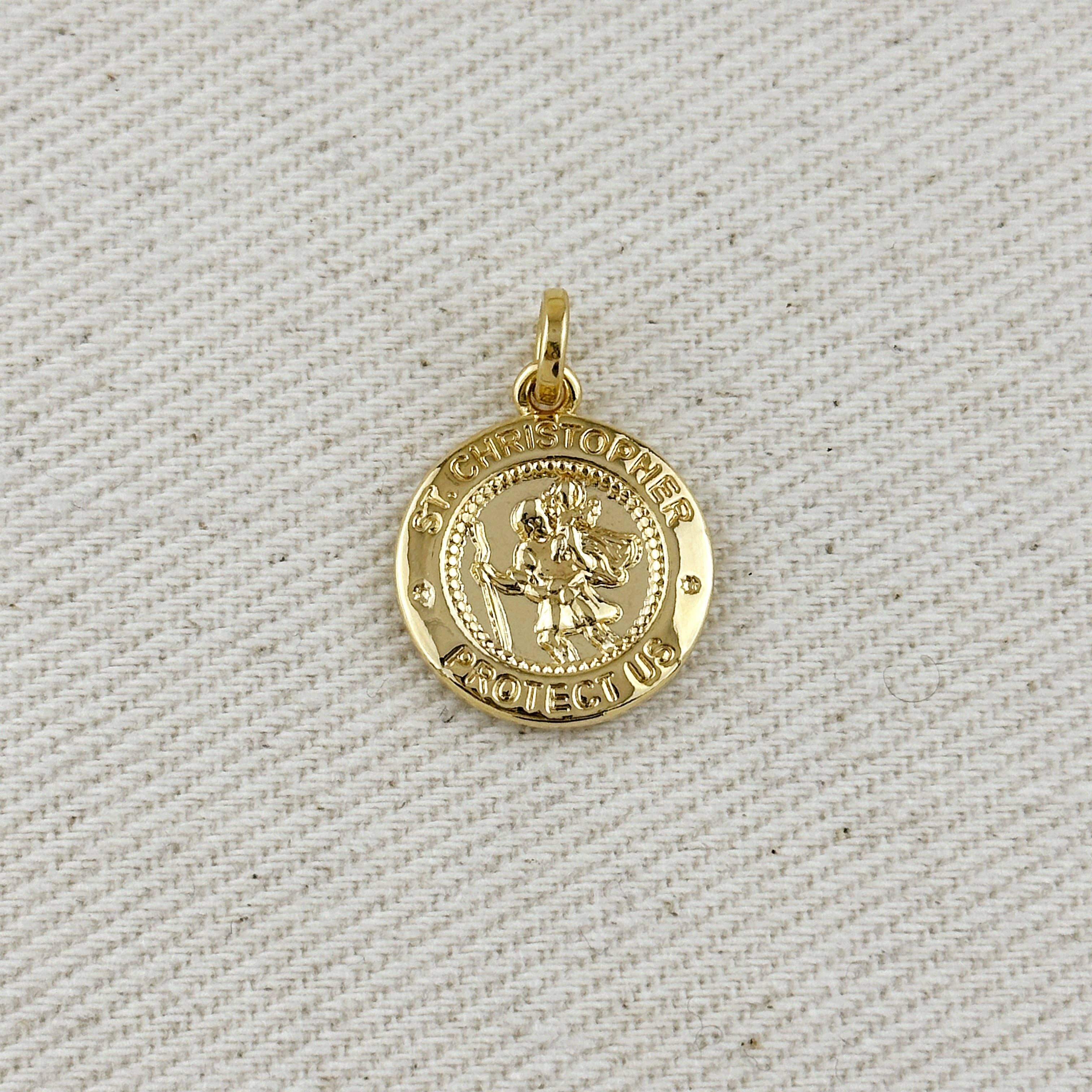 Gold pendant with 'St Christopher Protect Us' text on a textured beige background. Shop Rutledge Exchange for curated vintage and jewelry in Camden, SC.&nbsp;
