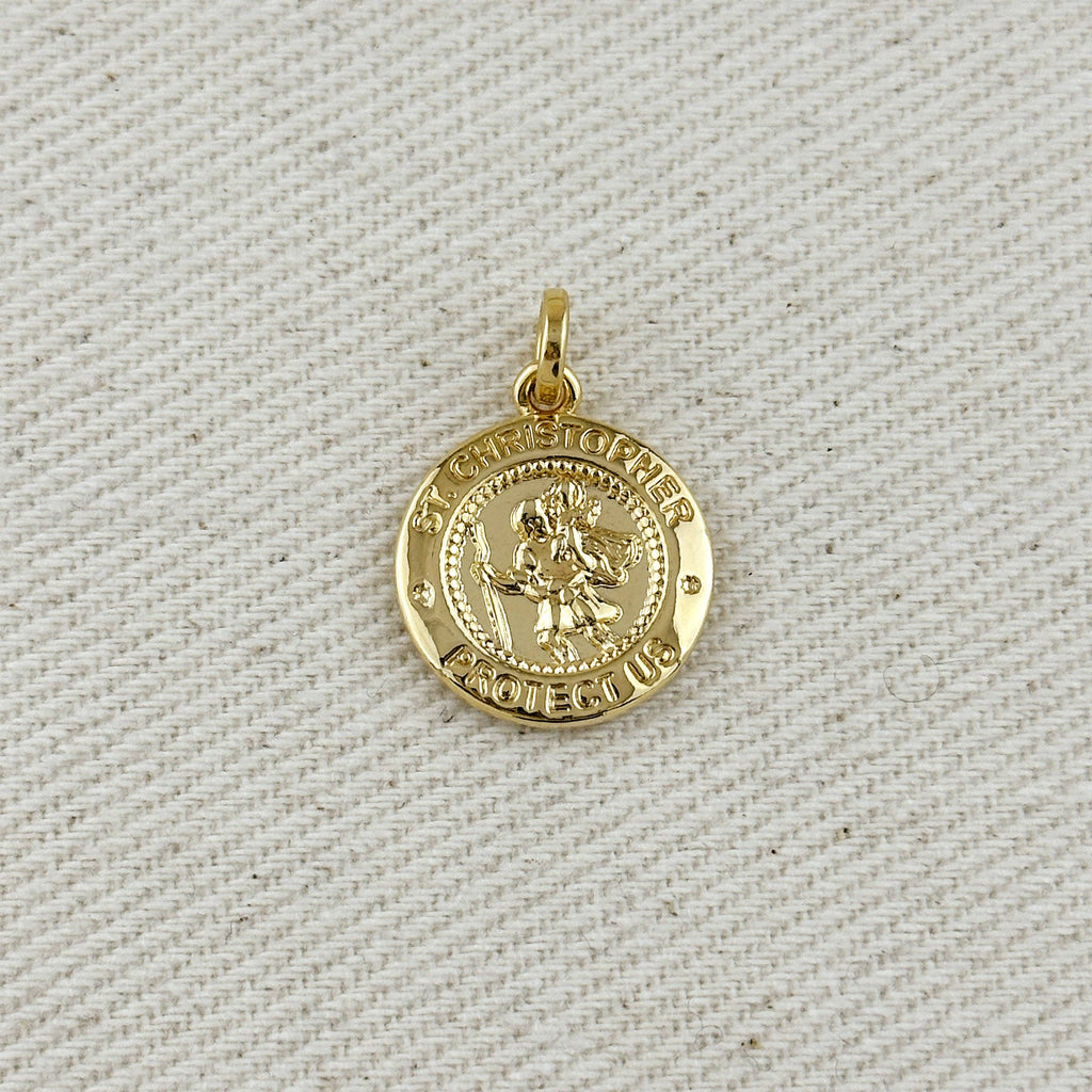 Gold pendant with 'St Christopher Protect Us' text on a textured beige background. Shop Rutledge Exchange for curated vintage and jewelry in Camden, SC.&nbsp;